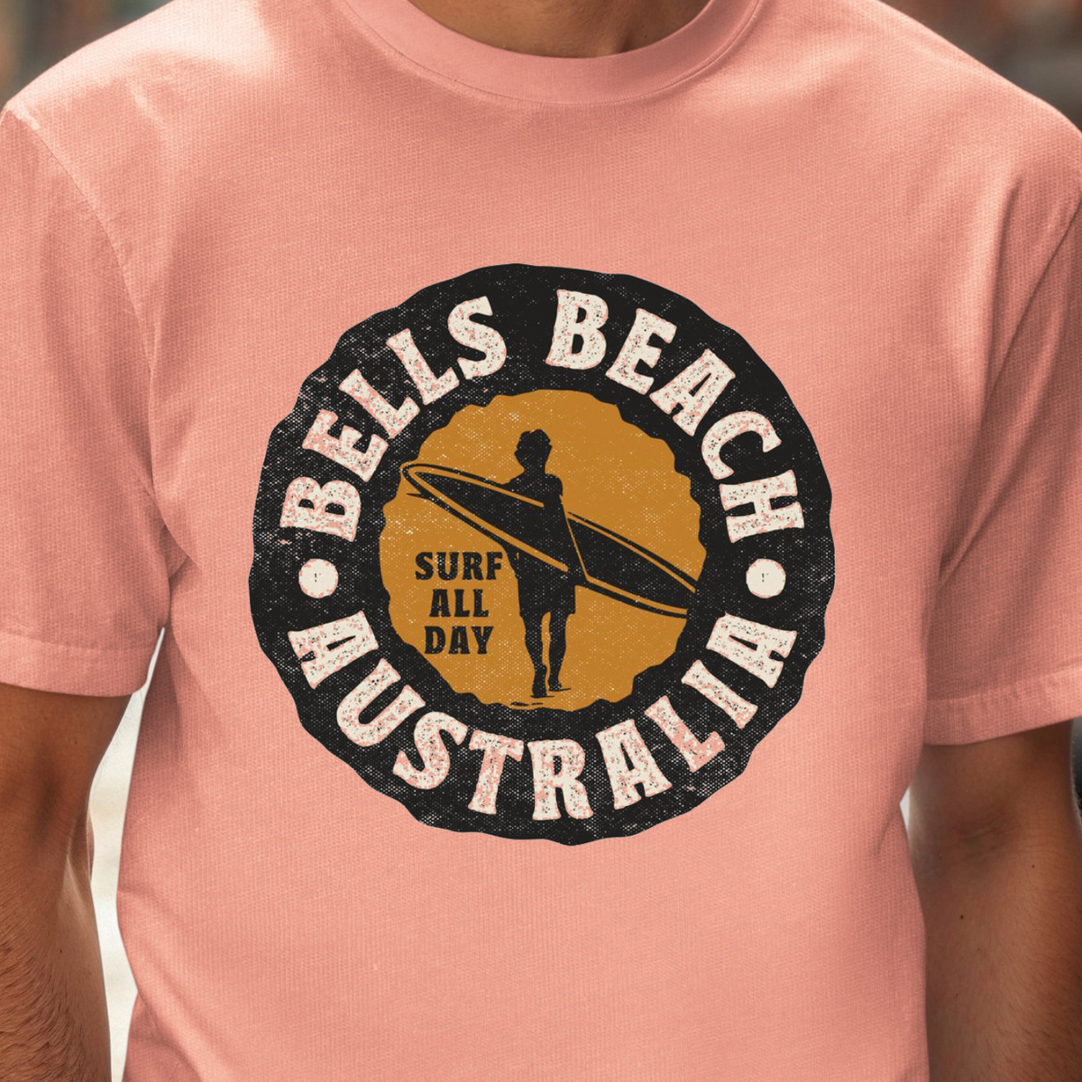 A man wearing a peach-pink t-shirt with a circular 'Bells Beach Australia Surf All Day' graphic on the chest, set against a blurred city street background.