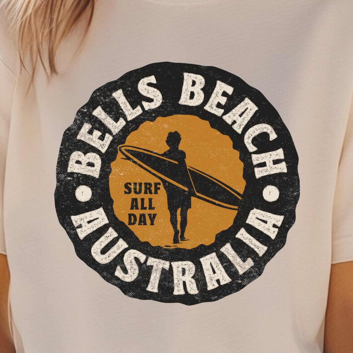 A woman wearing a cream-colored t-shirt with a distressed circular graphic that reads 'BELLS BEACH AUSTRALIA' and 'SURF ALL DAY' around a surfer silhouette, set against a blurred desert background.