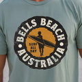 A man wearing a light blue-green t-shirt with a circular 'Bells Beach Australia Surf All Day' graphic on the chest, set against a blurred beach background.