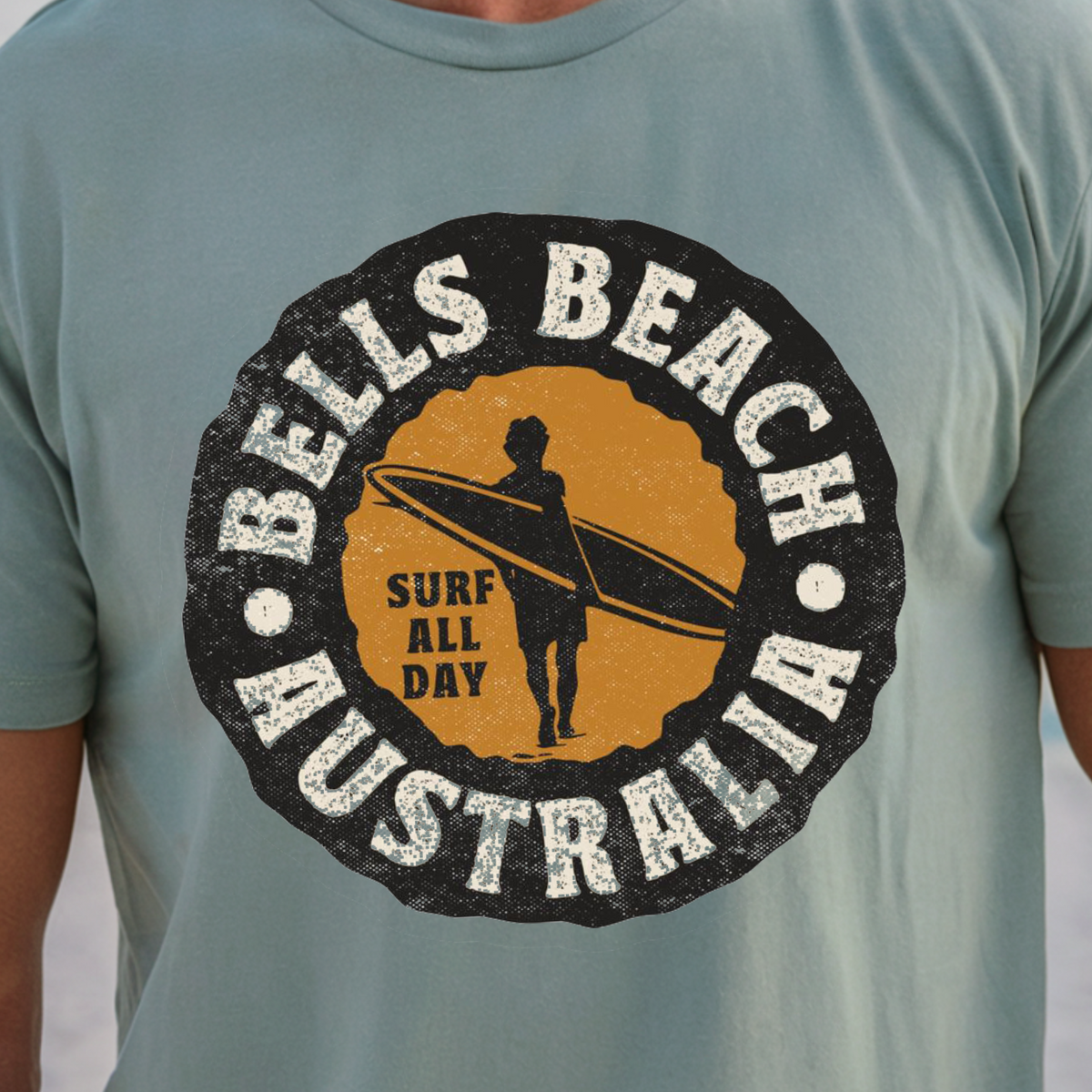 A man wearing a light blue-green t-shirt with a circular 'Bells Beach Australia Surf All Day' graphic on the chest, set against a blurred beach background.