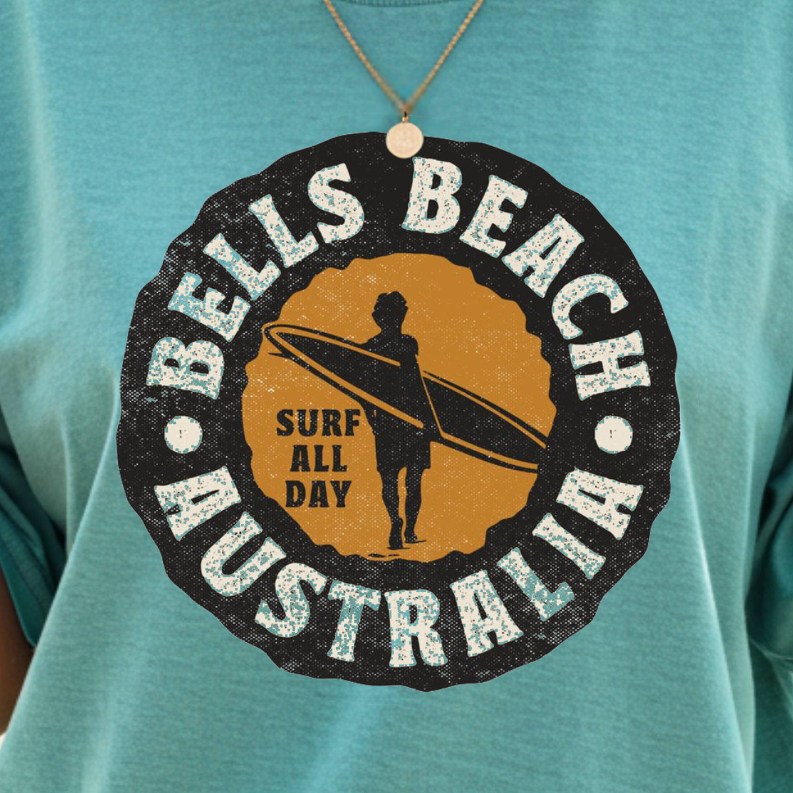 A woman on a beach wearing a teal t-shirt with a circular 'Bells Beach Australia Surf All Day' graphic in black and yellow. She is holding an iced coffee and wearing gold jewelry.