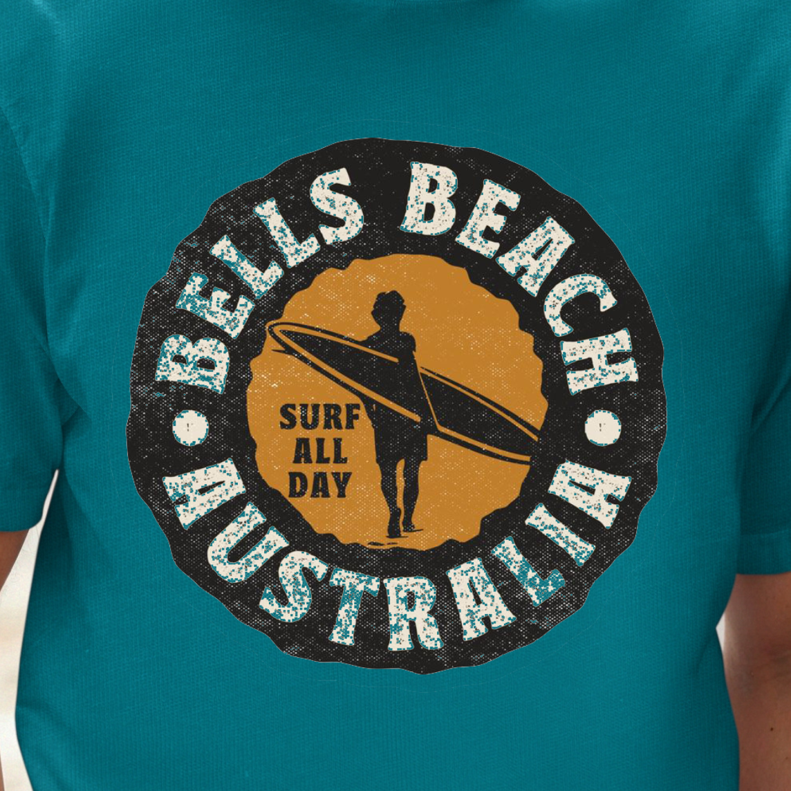 A man wearing a teal t-shirt with a circular 'Bells Beach Australia Surf All Day' graphic on the chest, set against a blurred city street background.