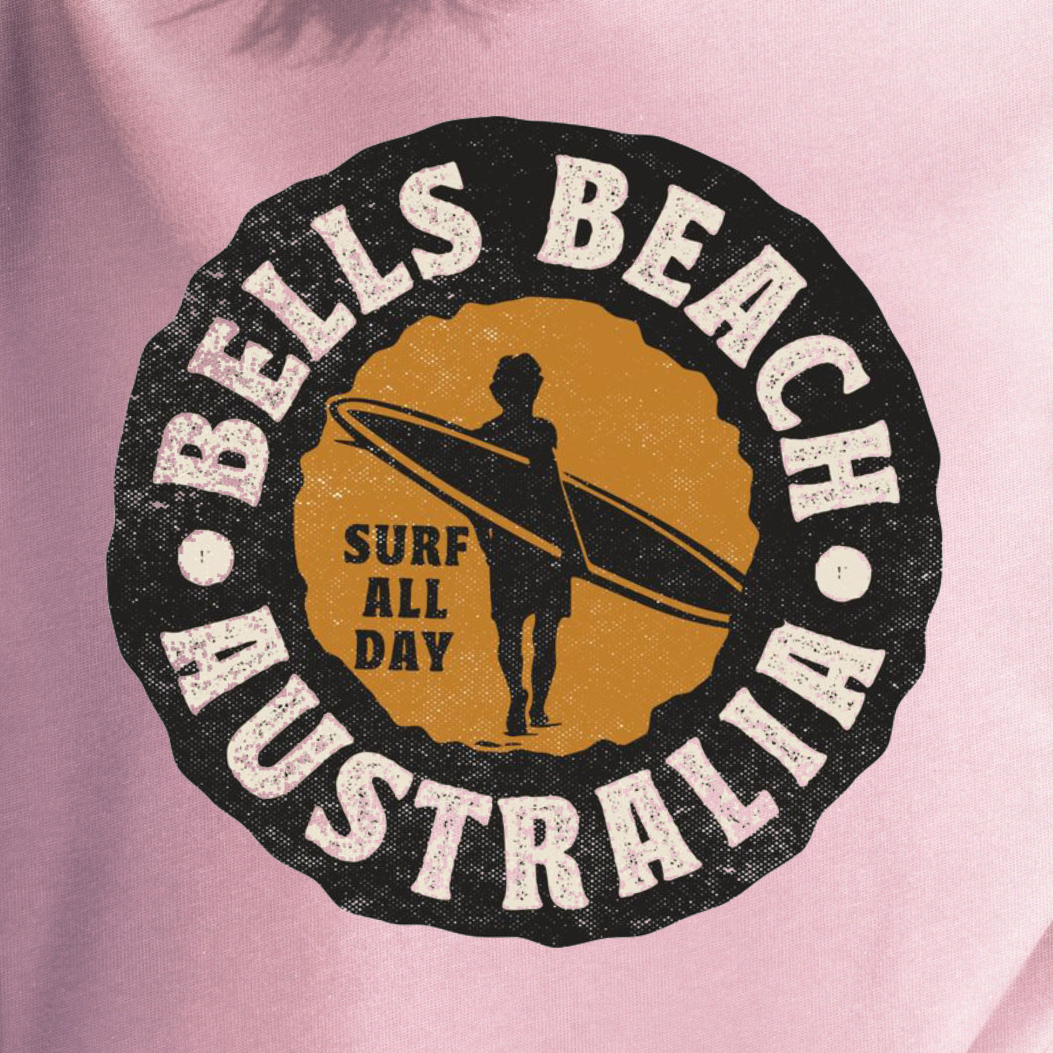 A woman wearing a light pink t-shirt with a circular 'Bells Beach Australia Surf All Day' graphic, standing on a beach with the ocean and blue sky in the background.