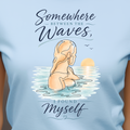 A woman wearing a light blue graphic t-shirt with a printed design of a woman meditating in the ocean waves with the text 'Somewhere Between the Waves, I Found Myself'.
