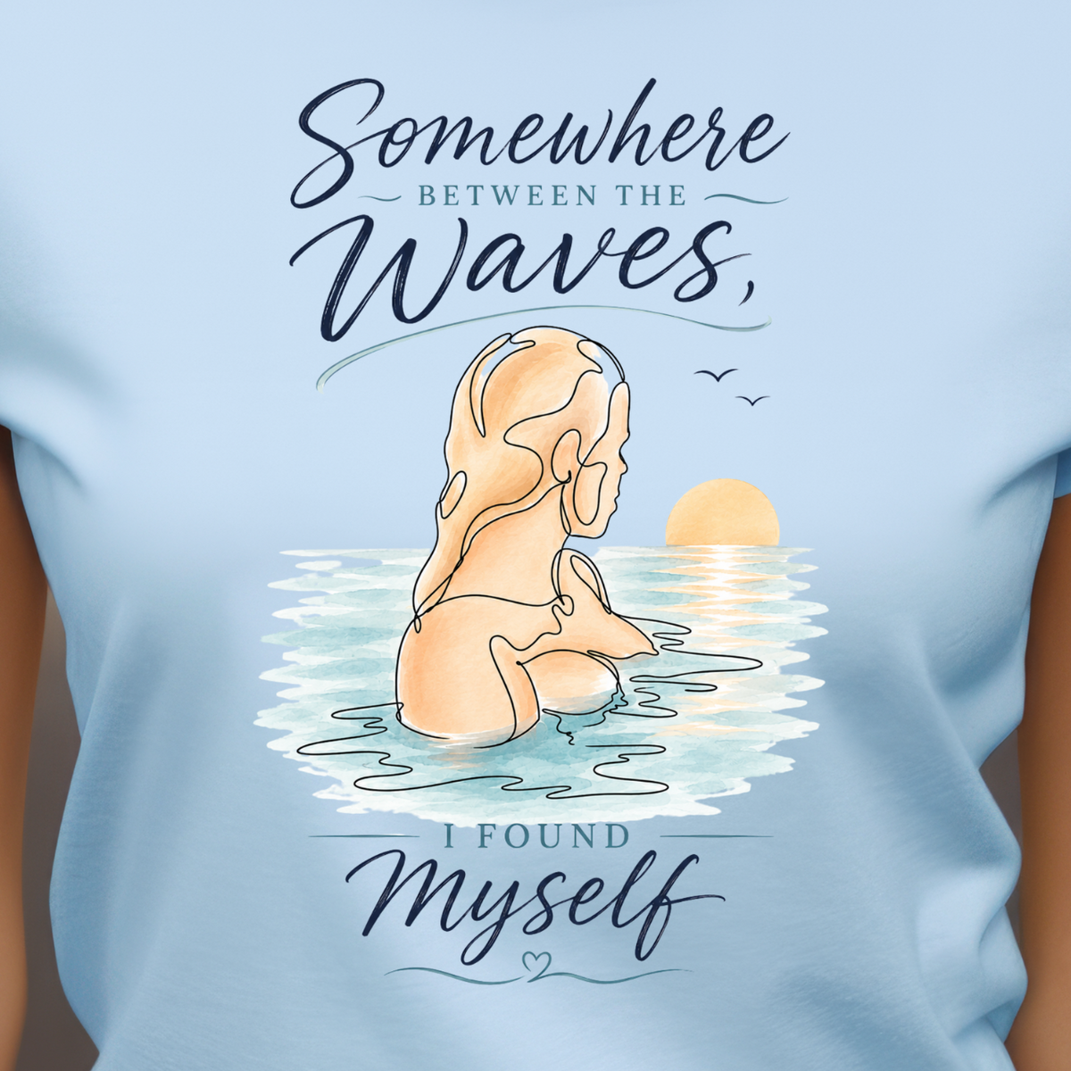 A woman wearing a light blue graphic t-shirt with a printed design of a woman meditating in the ocean waves with the text 'Somewhere Between the Waves, I Found Myself'.