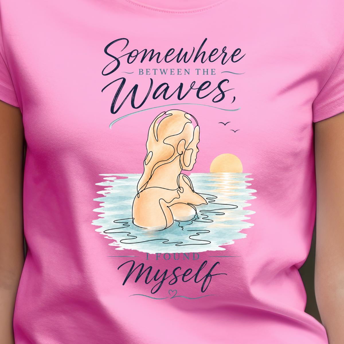 A woman wearing a bright pink short-sleeve t-shirt with a graphic print. The graphic features a woman meditating in the water with the text 'Somewhere between the waves, I found myself' in a dark purple script.