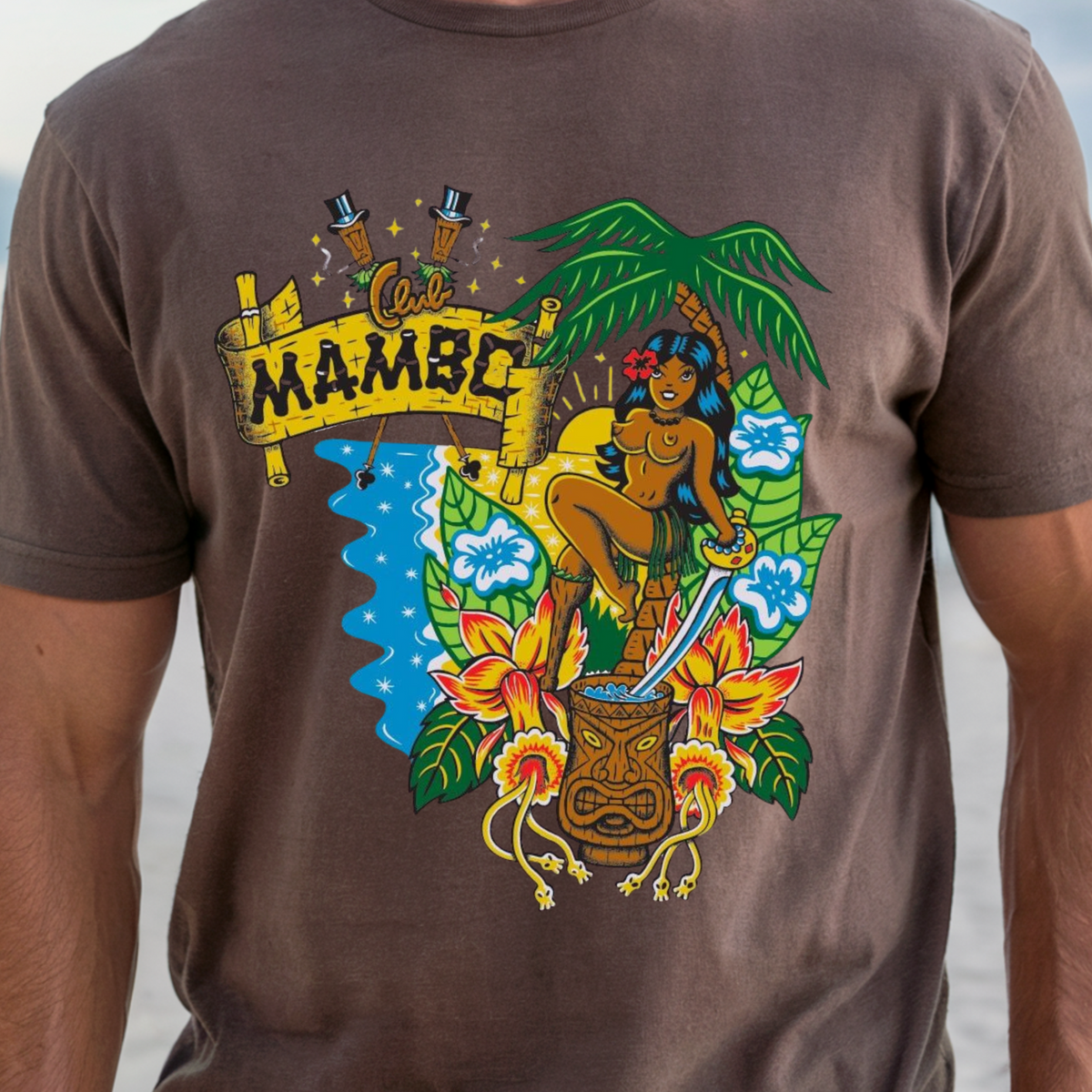 A man wearing a brown graphic t-shirt with a colorful Hawaiian-themed 'Get Mambo' print, standing on a sandy beach with the ocean in the background.