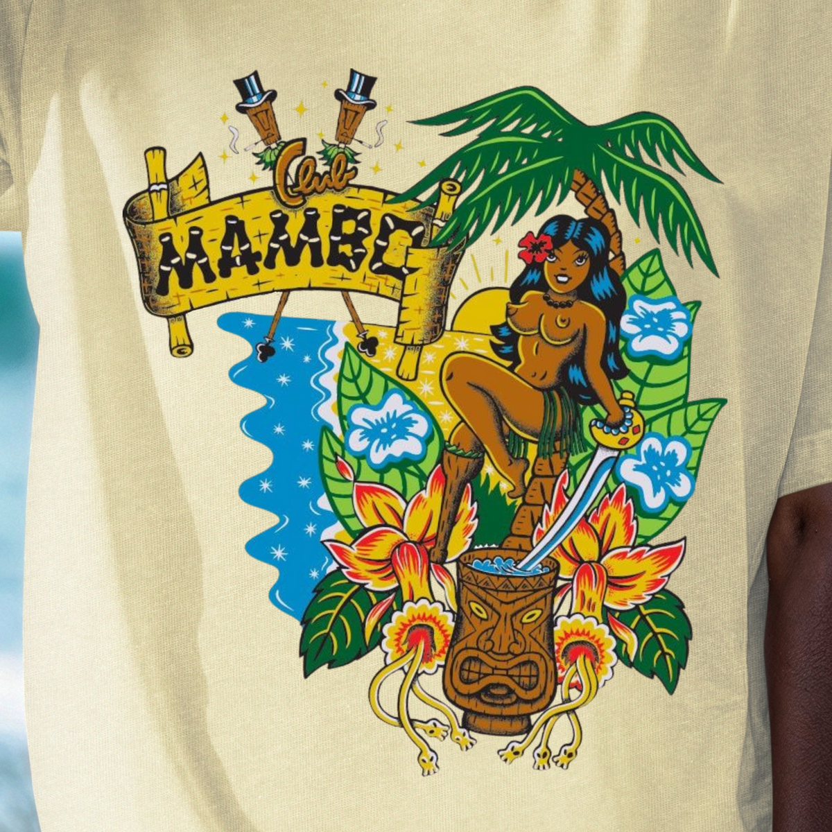 A person wearing a cream-colored graphic t-shirt with a colorful Hawaiian-themed print featuring the word 'MAMMY' on a banner, a tiki mug, flowers, and a palm tree, set against a blurred beach background.
