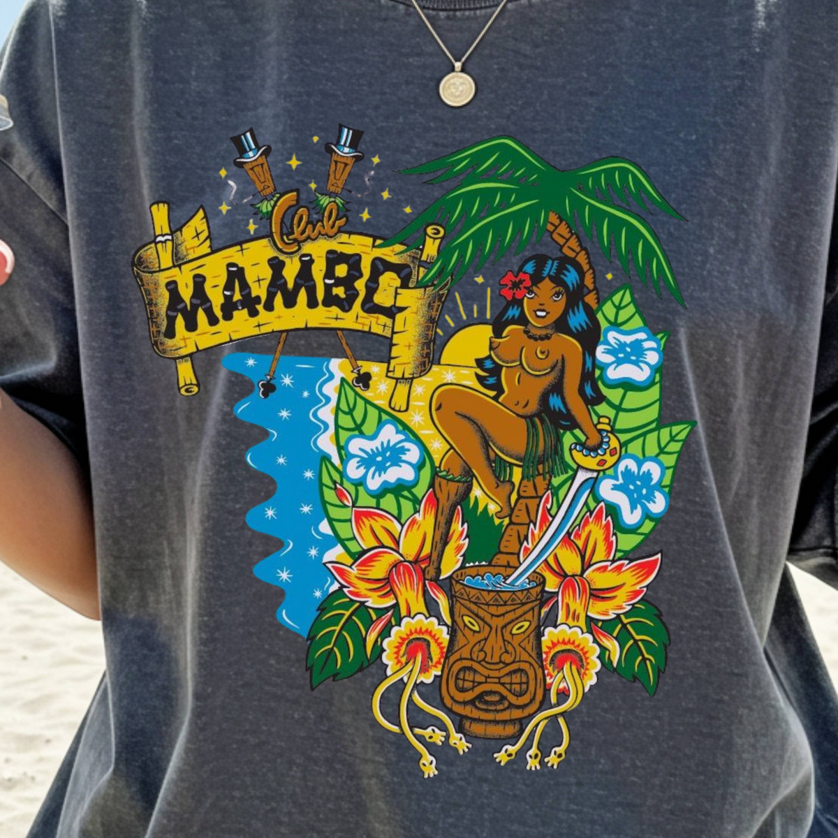 A person wearing an oversized charcoal gray graphic T-shirt on a beach, holding an iced coffee. The shirt features a colorful front print with a Hawaiian-style figure, flowers, a tiki mug, and a banner reading 'Oii MAMBO'.