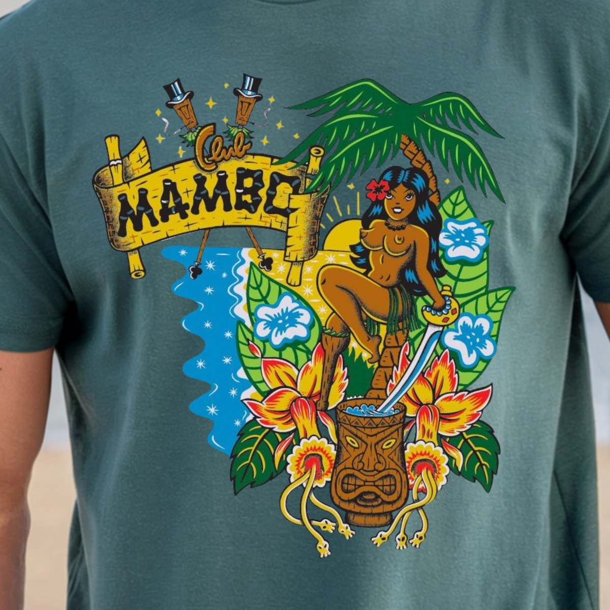A man wearing a teal green graphic t-shirt with a colorful Hawaiian-themed print featuring the word 'MAMBO' on a banner, a tiki mug, flowers, and a woman, set against a blurred beach and ocean background.