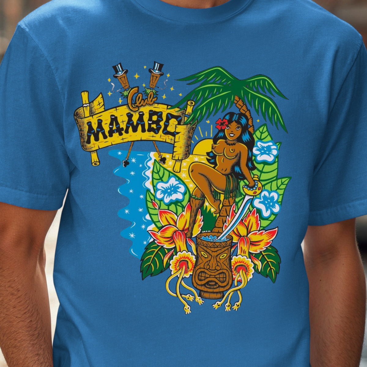 A man wearing a bright blue crewneck t-shirt with a colorful graphic print featuring a hula dancer, a tiki mug, hibiscus flowers, and a banner that reads 'MAMBO'.