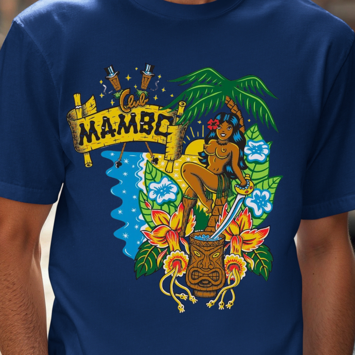 A man wearing a navy blue t-shirt with a colorful graphic print featuring a tiki, flowers, a palm tree, and a banner that reads 'MAMBO'. The background is a blurred street scene.