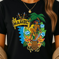 A woman wearing a black short-sleeve graphic T-shirt with a colorful tropical Mambo-themed print featuring a hula dancer, palm tree, flowers, and the word 'MAMBO'.