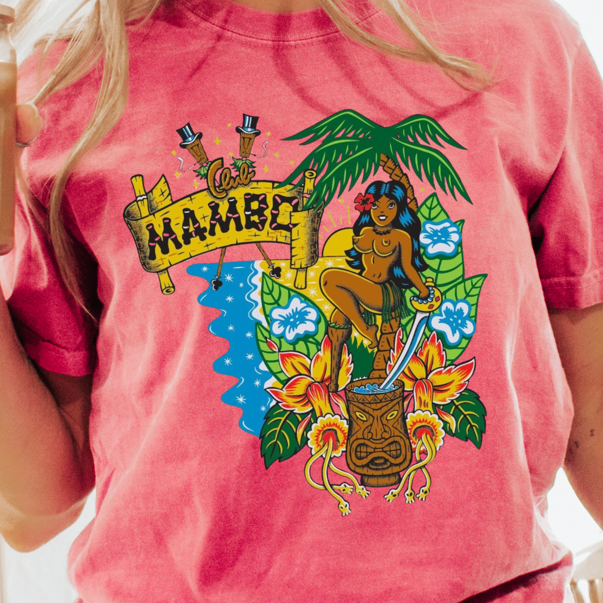 A woman wearing a pink mineral-washed graphic t-shirt with a colorful Hawaiian-themed print that reads 'Come HAMBONE'. She is holding an iced coffee in a bright indoor setting.