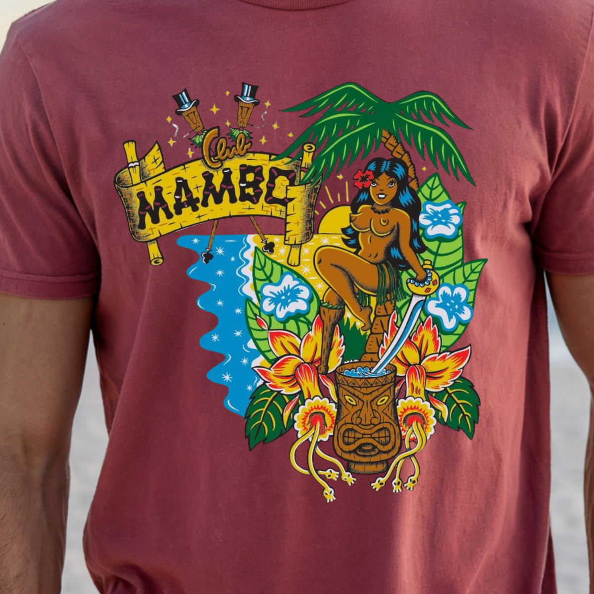 A man wearing a dusty rose short-sleeve t-shirt with a colorful graphic print on the front. The graphic features a tropical scene with a woman, a sword, a tiki mug, hibiscus flowers, a palm tree, and a banner reading 'MAMBC'.
