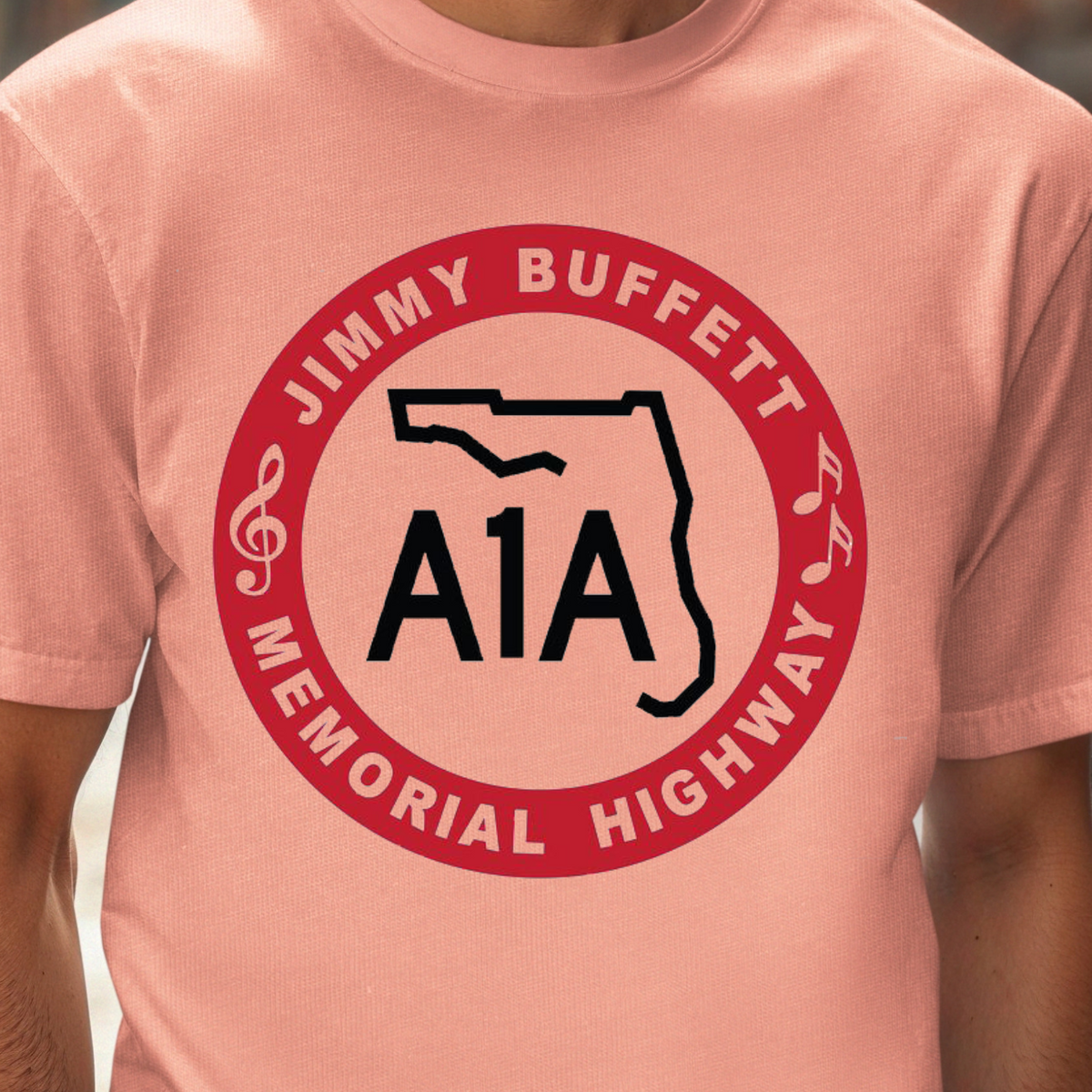 A man wearing a coral pink t-shirt with a red and black circular graphic that reads 'JIMMY BUFFETT MEMORIAL HIGHWAY' and 'A1A' with a Florida state outline.