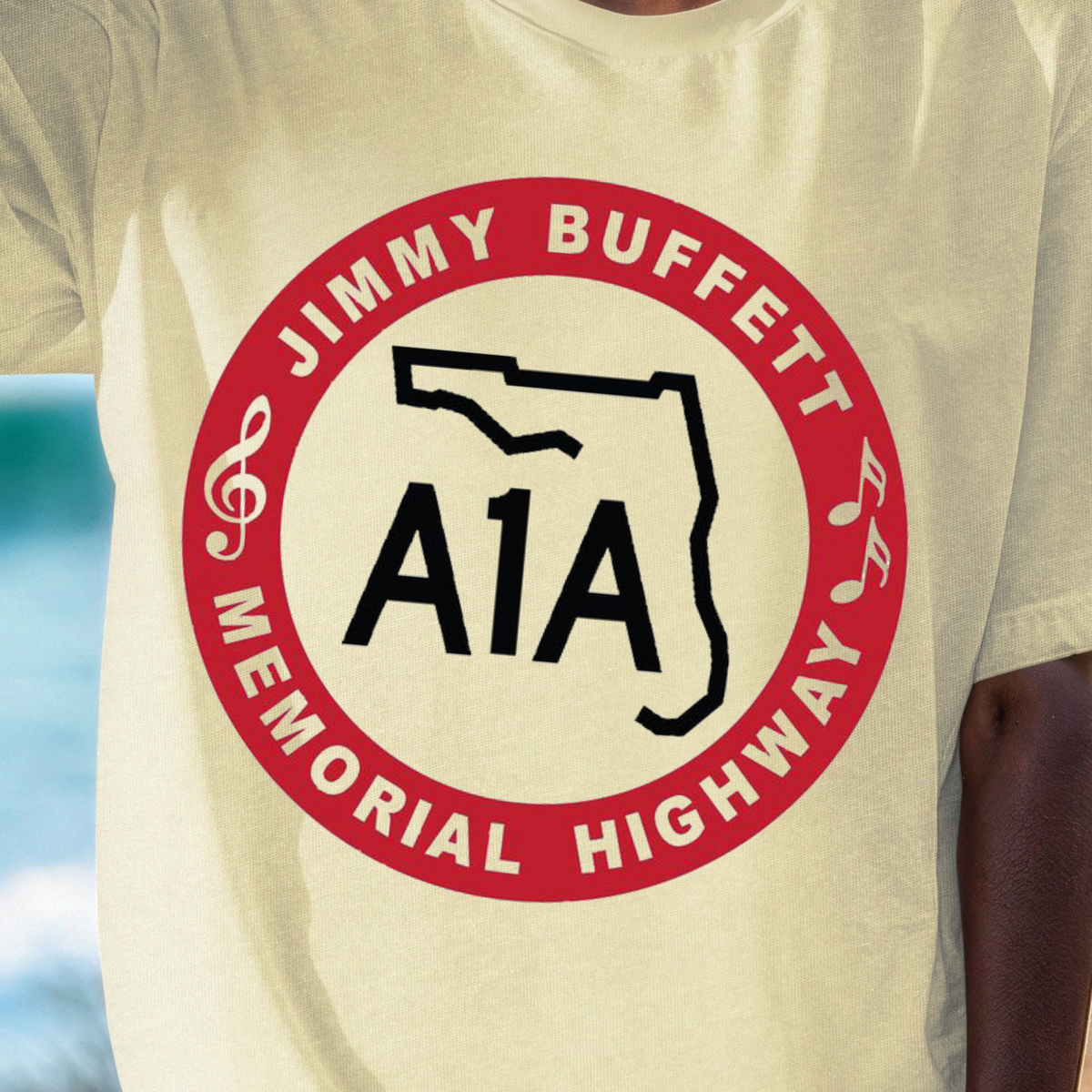 A person wearing a cream-colored t-shirt with a red and black graphic print that reads 'JIMMY BUFFETT A1A MEMORIAL HIGHWAY' with a Florida state outline, set against a blurred beach background.