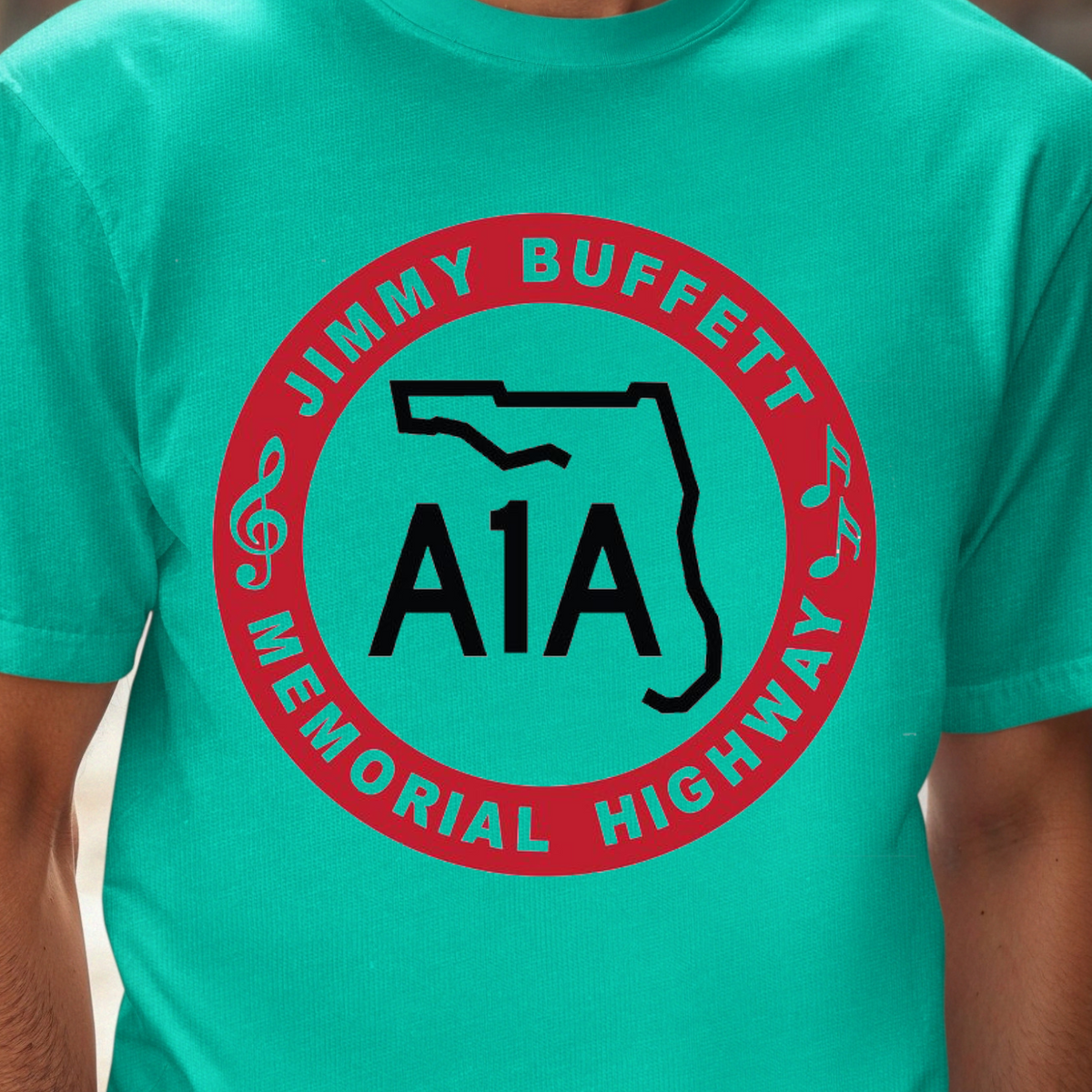 A man wearing a bright turquoise t-shirt with a circular red and black graphic on the chest that reads 'JIMMY BUFFETT MEMORIAL HIGHWAY A1A'. The background is a blurred outdoor street scene.