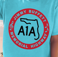 A person wearing a bright turquoise T-shirt with a large red and black circular graphic reading 'JIMMY BUFFETT A1A MEMORIAL HIGHWAY' and a black outline shape in the center, photographed outdoors near a beach.