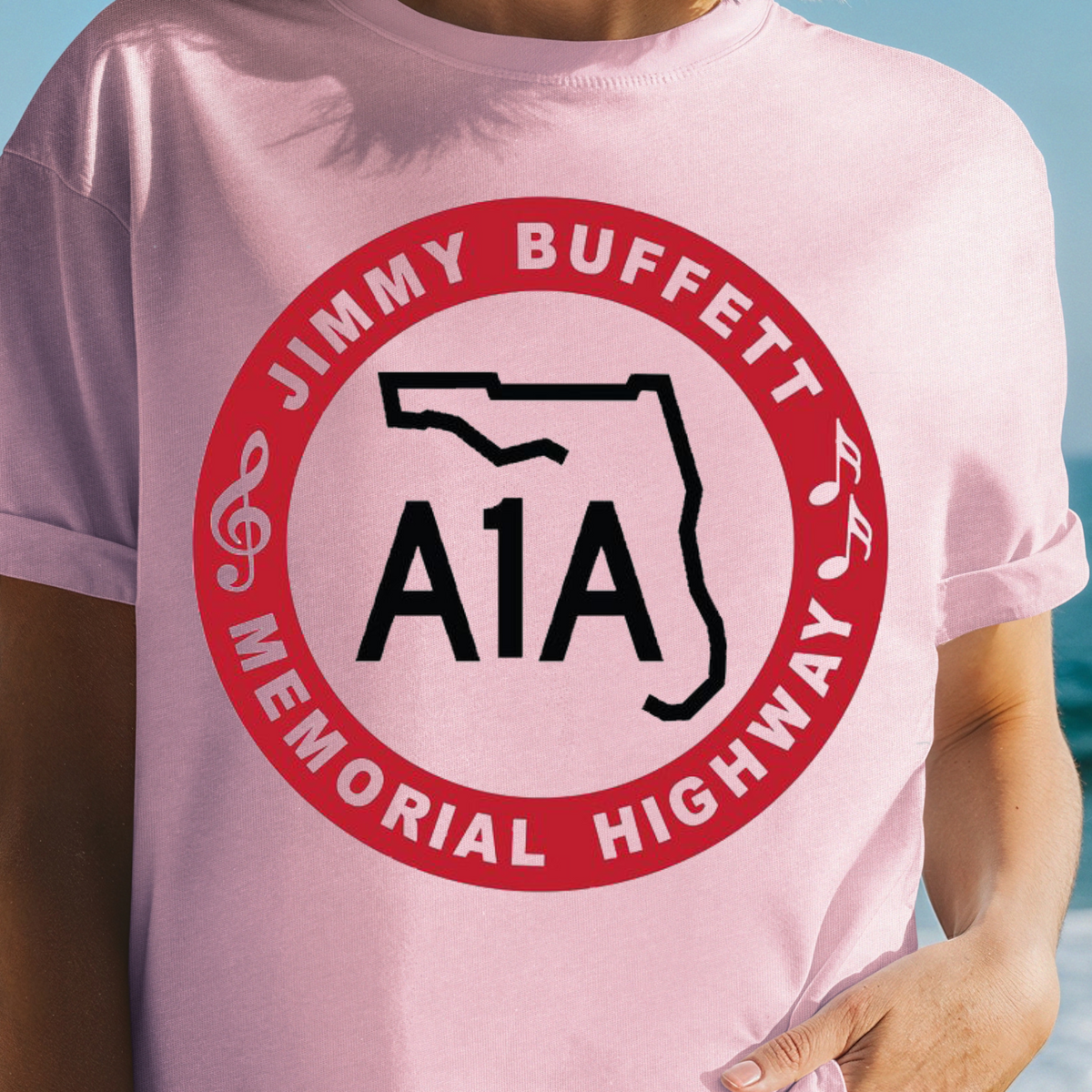 A woman wearing a light pink t-shirt with a red and black graphic that reads 'JIMMY BUFFETT MEMORIAL HIGHWAY A1A' on a beach background.