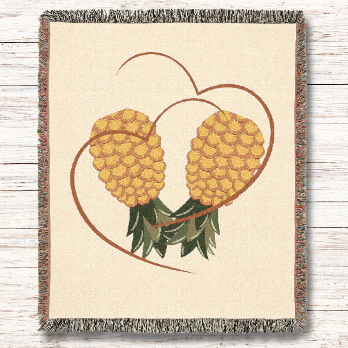 A rectangular woven throw blanket with fringed edges, featuring a design of two yellow pineapples inside a brown heart outline on a cream background, displayed flat on a light wood plank surface.