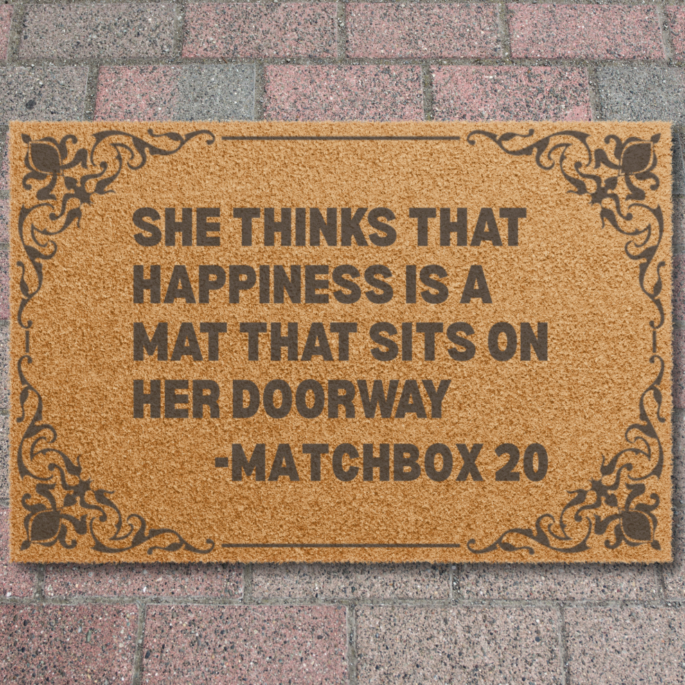 A rectangular natural coir doormat with black decorative corner flourishes and bold black text reading, 'SHE THINKS THAT HAPPINESS IS A MAT THAT SITS ON HER DOORWAY -MATCHBOX 20,' shown against a brick wall background.